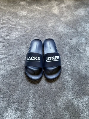 Marinblåa slides från Jack & Jones - Snygga marinblåa slides från Jack & Jones med vit logga på ovandelen. Tillverkade i syntetmaterial med räfflad sula för extra grepp. Perfekta för chill dagar hemma eller på stranden.