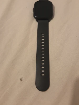 Svart smartklocka med laddare - Säljer en svart smartklocka med fyrkantig display och silikonarmband. Klockan kommer med en rund magnetisk laddare som ansluts via kabel. Perfekt för dig som vill ha koll på notiser och hälsa direkt på handleden.