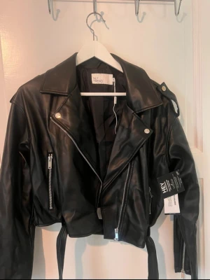 Svart skinnjacka från NLY Trend - Snygg svart skinnjacka från NLY Trend med klassisk bikerstil, silvriga dragkedjor och knappar. Jackan har bred krage, bälte i midjan och två fickor med dragkedja. Perfekt för dig som vill ha en edgy look.