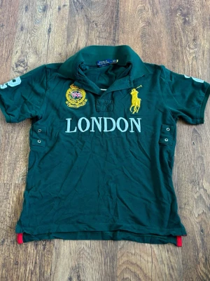Grön ”Chief Keef”  ”LONDON” Polo från Ralph Lauren - (OBS Det står storlek L men har krympt till närmare S!) Mörkgrön ”Chief Keef” pikétröja från Polo Ralph Lauren med stor gul broderad logga, texten 'LONDON' framtill och patch med brittisk flagga. Klassisk krage, korta ärmar och knappdetaljer i sidan. Perfekt för en sportig och stilren look.