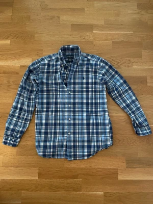Blårutig skjorta från Ralph Lauren - Klassisk blårutig skjorta från Ralph Lauren med lång ärm och knappar framtill. Skjortan har den ikoniska broderade loggan på bröstet och är tillverkad i mjuk bomull. Perfekt för en avslappnad men ändå stilren look.