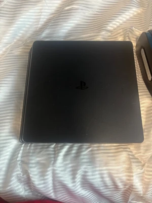 PlayStation 4 Slim svart konsol - Säljer en svart PlayStation 4 Slim från Sony. Konsolen har en stilren, matt finish och den klassiska PlayStation-loggan på ovansidan. Formen är rektangulär med rundade hörn och konsolen är tillverkad i plast. Perfekt för gaming och streaming.