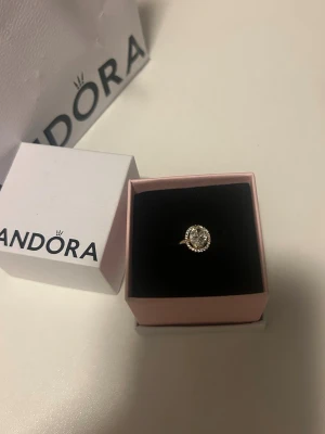 Guldfärgad ring med stenar från Pandora - Snygg guldfärgad ring från Pandora med en stor rund klar sten i mitten, omgiven av mindre gnistrande stenar. Ringen har en klassisk och elegant design som verkligen sticker ut. Perfekt accessoar för dig som gillar lite extra bling. Storlek 52 ALE MET 52. Kommer med påse och ringhållaren
