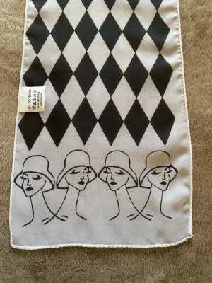 Svartvit smal sjal med grafiskt mönster - Smal sjal i svart och vitt  med grafiskt harlequinmönster och stiliserade ansikten med hattar längs kanterna. Tillverkad i 100% polyester, perfekt att knyta runt halsen eller i håret för en cool look.