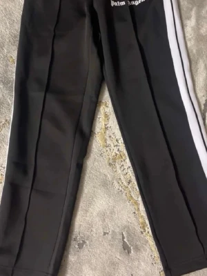 Palm Angels svarta mjukisbyxor - Svarta mjukisbyxor från Palm Angels med vita sidoränder och elastisk midja. Byxorna har raka ben och logga tryckt framtill. Perfekta för en avslappnad och trendig streetwear-look.