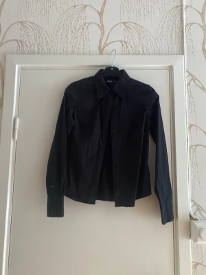 Svart långärmad skjorta från Weekday - Stilren svart skjorta från Sisters med klassisk krage och knäppning framtill. Skjortan har lång ärm och är tillverkad i ett slätt bomullstyg. Perfekt för en clean och enkel look.