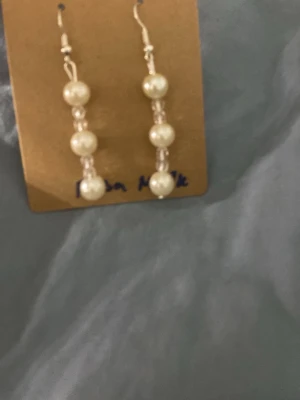 Hängande örhängen med pärlor - Snygga hängande örhängen med tre vita pärlor och små genomskinliga pärldetaljer mellan varje. Örhängena har krokar i metall och en stilren design som passar till många olika looks. Perfekt accessoar för dig som gillar klassiska smycken med en modern touch.