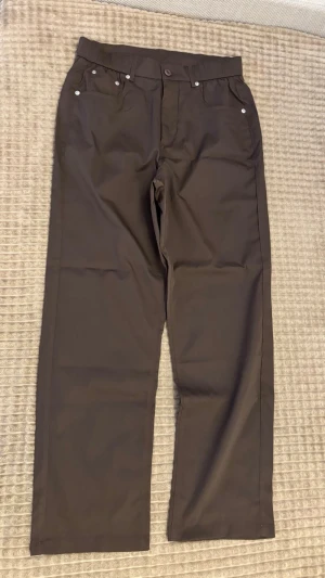 Bruna raka chinos med fickor - Snygga bruna chinos med raka ben och klassisk passform. Byxorna har framfickor med nitar och två bakfickor. Perfekta för en stilren look och tillverkade i ett mjukt bomullsmaterial. Bälteshällor upptill för extra stilpoäng.