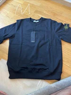 Svart sweatshirt från Stone Island - Snygg svart sweatshirt från Stone Island med klassisk rund hals och ribbade muddar. Tröjan har den ikoniska loggan på vänster ärm och är tillverkad i mjukt bomullsmaterial. Perfekt för en clean och stilren look.