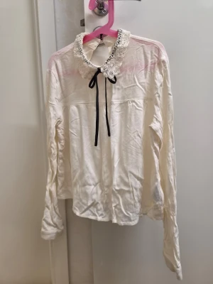 Vit blus med spetskrage och rosett H&M 34 - Söt vit blus med lång ärm, spetskrage och svart rosett framtill. Blusen har en feminin och romantisk stil med dekorativ spets runt kragen och en liten öppning i nacken. 