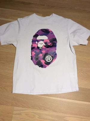 BAPE vit t-shirt med camo-print - Vit t-shirt från BAPE med ikoniskt ape head-tryck i lila, rosa och svart camo på både fram- och baksida. Klassisk passform och rund hals. Tillverkad i mjuk bomull, perfekt för en streetwear-look. 
