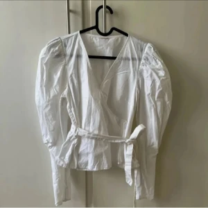 Vit omlottblus med puffärm och knyt - Säljer en vit omlottblus med långa puffärmar och knyt i midjan. Blusen har v-ringning och är tillverkad i ett tunt, lätt material som ger en luftig känsla. Perfekt för dig som gillar romantiska och trendiga detaljer. Minns inte men den är köpt på Zara eller hm 