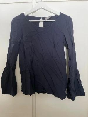Mörkblå blus från Vero Moda - Snygg mörkblå blus från Vero Moda med v-ringning och långa, lätt utsvängda ärmar. Blusen har en enkel men trendig design och är tillverkad i ett mjukt, tunt tyg som ger en avslappnad look. Perfekt att styla med jeans eller kjol.