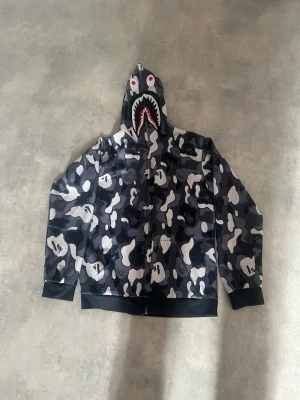 BAPE camo hoodie med hajmotiv - Säljer en svart, grå och vit camo hoodie från BAPE med hajmotiv på huvan och dragkedja hela vägen upp. Huvan har broderade detaljer med ögon och tänder samt texten WGM på sidan. Perfekt för dig som gillar streetwear och vill sticka ut.