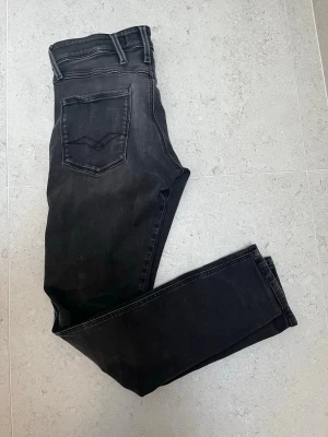 Svarta anbass jeans från Replay - Snygga svarta slim fit jeans från Replay med klassisk femficksdesign och diskreta slitningar. Jeansen har en smal passform och är tillverkade i stretchigt material för extra komfort. Perfekta till en avslappnad streetstyle-look. Storlek 30/32.