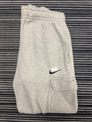 Grå mjukisbyxor från Nike - Snygga grå cargobyxor från Nike med elastisk midja och stora fickor på sidorna. Byxorna har en klassisk Nike-logga på ena benet och är gjorda i mjuk bomull. Perfekta för en avslappnad och sportig look.