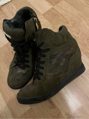 Olivgröna camo high-top sneakers - Snygga olivgröna high-top sneakers från Guess med camo-mönster på sidorna och svarta skosnören. Skorna har en robust design med mockadetaljer och en svart sula. Perfekta för dig som vill ha en cool streetstyle-look.
