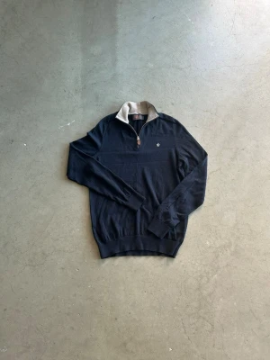 Morris quarterzip - Mörkblå långärmad tröja med halvzip och beige krage. Tröjan har en liten broderad logga på bröstet och ribbade muddar vid ärmslut och nederkant. Perfekt för lager-på-lager och snygg till jeans. Har en defekt vid vänster armbåge! Skriv vid frågor