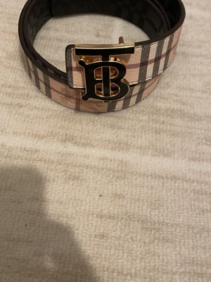Rutig Burberry skärp med guldspänne - Säljer ett klassiskt Burberry skärp med det ikoniska rutiga mönstret i beige, brunt och svart. Skärpet har ett stort guldigt TB-spänne framtill och är gjort i skinn. Perfekt accessoar för att lyfta vilken outfit som helst.