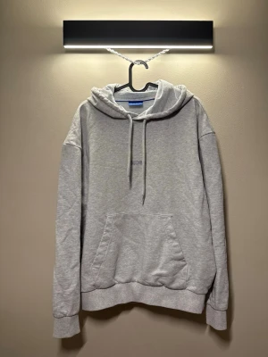 Grå hoodie från Hugo med logga - Snygg grå hoodie från Hugo med diskret broderad logga på bröstet. Hoodien har en klassisk känguruficka, dragsko i huvan och ribbade muddar vid ärmar och nederkant. Tillverkad i mjuk bomullsblandning som känns skön mot huden. Blå ”HUGO” på baksidan.