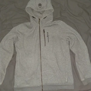 Sailracing zip hoodie - Ljusgrå hoodiejacka från Sail Racing med hel dragkedja, bröstficka med dragkedja och broderad logga på bröstet och huvan. Storlek 170 finns nån fläck på tröjan