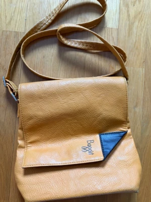 Gul axelväska från Baggit - Snygg gul axelväska från Bagotti i konstläder med justerbar axelrem. Väskan har ett lock med svart detalj och Bagotti-logga, samt en praktisk dragkedjeficka på baksidan. Perfekt för dig som vill ha en unik och trendig accessoar.