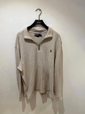 Beige halfzip från Polo Ralph Lauren - Snygg beige halfzip-tröja från Polo Ralph Lauren med klassisk blå logga broderad på bröstet. Tröjan har hög krage, dragkedja med läderdetalj och är tillverkad i mjukt bomullsmaterial. Perfekt för en clean och avslappnad stil.