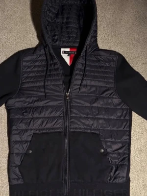 Svart hoodie från Tommy Hilfiger - Snygg svart hoodie från Tommy Hilfiger med quiltad framsida och huva. Dragkedja hela vägen, två stora fickor framtill och diskret logga på mudden. Ärmar och fickor i mjukt tyg, medan framsidan och huvan är lätt vadderade för extra stil.