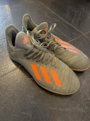 Adidas grå-orange fotbollsskor - Säljer ett par grå fotbollsskor från Adidas med klassiska orangea stripes och detaljer. Skorna har snörning, låg profil och ovandel i syntetmaterial för bra bollkontroll. Snygg design som sticker ut på planen.