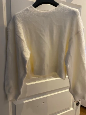Vit fluffig cropped stickad tröja - Supermjuk och fluffig vit stickad tröja med croppad passform och rund hals. Perfekt för lager på lager och ger en trendig look. Långa ärmar och enkel design som passar till det mesta.