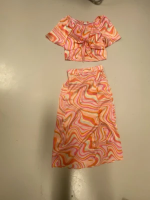 Färgglatt set med swirlmönster - Säljer ett tvådelat set med kort topp och kjol i ett färgglatt swirlmönster i rosa, orange och vitt. Toppen har knytning framtill och kjolen är midilång med slits. Materialet känns lätt och mjukt, perfekt för dig som gillar retrovibes och att sticka ut.