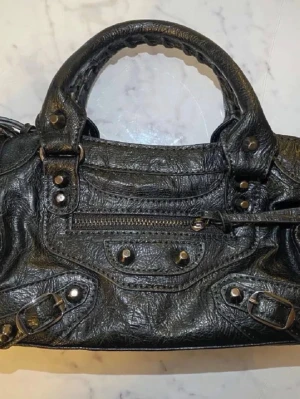 Svart Balenciaga City handväska - Cool svart Balenciaga City dupe handväska i skinn med klassiska nitar och dragkedjedetaljer. Väskan har både handtag och avtagbar axelrem, samt flera fack och dekorativa spännen. Perfekt för dig som gillar edgy och tidlös design. ❣️❣️❣️❣️