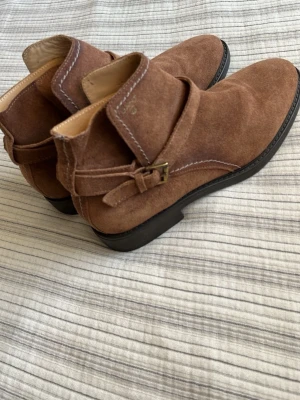 Bruna mocka stövletter från Gant - Snygga bruna stövletter från Gant i mjuk mocka med dekorativt spänne på sidan. Skorna har låg klack, rundad tå och kontrastsömmar som ger en stilren look. Perfekta för dig som gillar klassisk men trendig stil. Superbekväma och bra i storleken, nyskick!