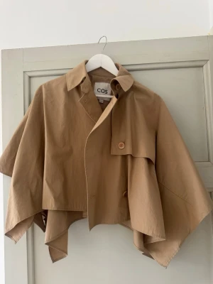 Kort beige trenchcoat från COS - Säljer en beige trenchcoat från COS med kort, asymmetrisk passform och klassisk krage. 