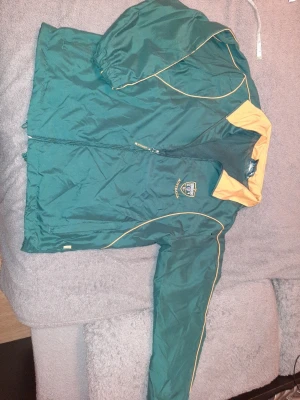 Australia Rugby Team trainingjacka från England Rugby Sevens, år 2010s - Retro snygg grön trainingjacka med gul insida på kragen och tunna gula linjer längs ärmar och bröst. Jackan har dragkedja framtill, resår i ärmslut och en broderad logga på bröstet. Perfekt för utomhusaktiviteter och sport. Materialet är lätt och vindtätt. Limited edition from 2010s' game. UNISEX. Storlek S för man,  storlek M för kvinna. 