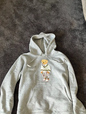 Grå hoodie Polo Bear Ralph Lauren - Säljer en grå hoodie från Ralph Lauren med den ikoniska Polo Bear-trycket på bröstet. Hoodien har huva med dragsko och en stor magficka. Materialet är mjukt och skönt
