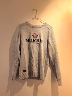 Grå stickad tröja från Morris - Snygg grå stickad tröja från Morris med Union Jack-logga och texten 'Morris Since 1968' broderat på bröstet. Tröjan har rund halsringning, långa ärmar och ribbade muddar vid ärmslut och nederkant. Perfekt för en stilren vardagslook.