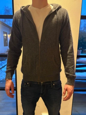 Grå hoodie i kashmir med dragkedja - Mysig grå hoodie från DAVIDA i mjuk kashmir. Hoodien har hel dragkedja framtill, två fickor och huva med snören. Perfekt för dig som gillar en enkel men stilren look. Passformen är normal och ärmarna är långa.