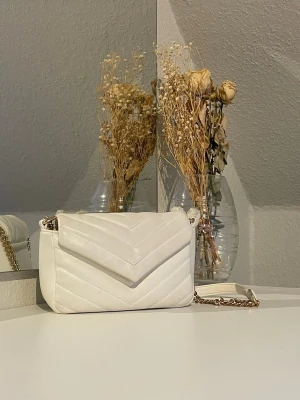 Vit handväska med guld kedja - Snygg vit handväska i quiltat mönster med V-formade sömmar. Väskan har en guldfärgad kedjerem och en magnet som håller väskan stängd💕