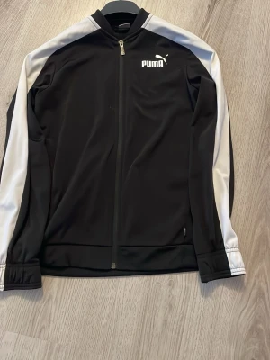 Svart träningsjacka från Puma - Säljer en svart träningsjacka från Puma med vita detaljer längs ärmarna och dragkedja framtill. Jackan har ståkrage och Puma-logga på bröstet. Perfekt för träning eller chill. Materialet är mjukt och stretchigt för bästa komfort.