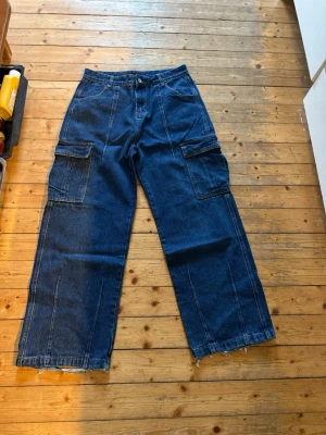 Blå cargojeans med vida ben - Säljer ett par blå cargojeans med bred passform och stora fickor på sidorna. Jeansen har råa, fransiga benslut och klassisk femficksdesign. Perfekta för en avslappnad streatwear. Pris kan diskuteras 