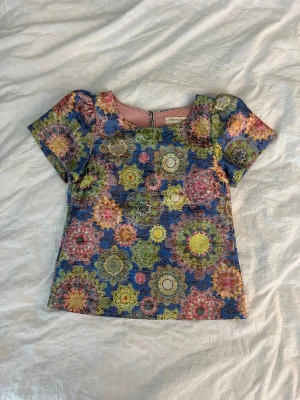 Färgglad blommig topp - Kortärmad topp från traffic people med färgglatt blommigt+glittrigt mönster i blått, rosa, gult och grönt. Toppen har rund halsringning och en lätt strukturerad yta. Perfekt för dig som gillar statement-plagg och vill sticka ut.