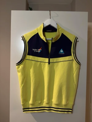 Vintage sportväst från Le Coq Sportif - Gul ärmlös sportväst från Le Coq Sportif med marinblå detaljer och dragkedja framtill. Västen har broderade loggor och text på bröstet samt ribbade muddar med blå ränder vid ärm och nederkant. Tillverkad i ett ribbat material som känns sportigt och retro.