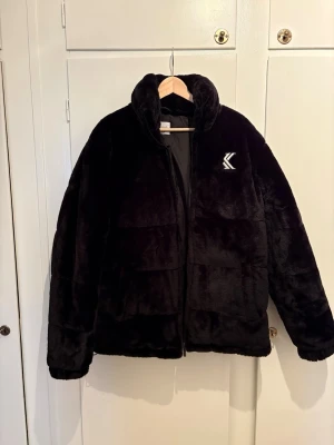 Svart pufferjacka från Karl Kani - Säljer en svart pufferjacka från Karl Kani med mjuk, fluffig yta och hög krage. Jackan har broderad logga fram och stor Karl Kani-text på ryggen. Perfekt för kalla dagar och riktigt snygg streetstil. Pris går att diskutera.