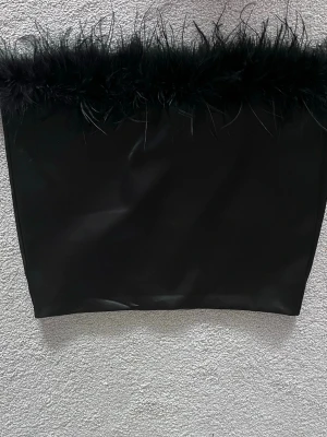 Svart kortkjol med fjädrar - En svart kortkjol med en cool fjäderdetalj längs överkanten. Kjolen har en slät och glansig yta som ger en snygg look. Perfekt för dig som vill sticka ut och addera lite edge till din outfit.