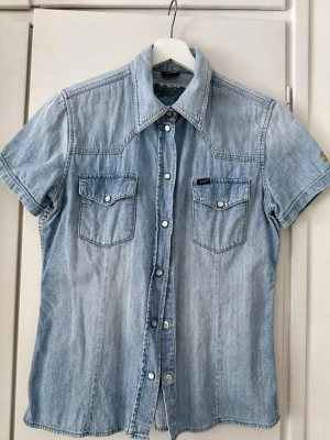 Ljusblå jeansskjorta från Lee - Säljer en klassisk ljusblå jeansskjorta från Lee med korta ärmar, tryckknappar och två bröstfickor. Skjortan har en snygg western-inspirerad design och är gjord i bomull. Perfekt för en avslappnad och cool stil.