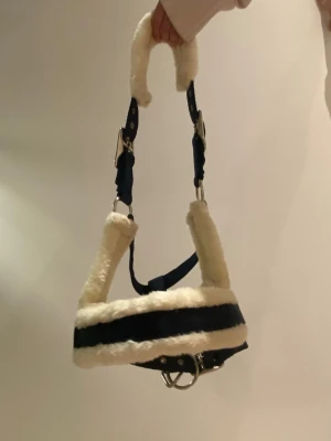 Marinblå grimma med fuskpäls - Säljer en marinblå grimma med mjuk vit fuskpäls och silvriga spännen. Grimman har justerbara remmar och vadderade partier för extra komfort. Perfekt till häst eller ponny och ger en snygg look i stallet. Använt max 2 gånger