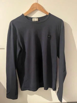 Mörkblå långärmad t-shirt Acne Studios - Snygg mörkblå långärmad t-shirt från Acne Studios med klassisk rund hals och den ikoniska lilla patchen på bröstet. Tillverkad i mjuk bomull för en skön känsla och clean look. Perfekt för dig som gillar minimalistisk stil.