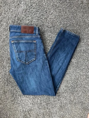 Blå slim fit jeans från tiger of sweden - Säljer ett par klassiska blå slim fit jeans från Studios. Jeansen har fem fickor, normal midja och raka ben. Tillverkade i mjuk denim med snygga kontrastsömmar och läderpatch bak i midjan. Perfekta till en avslappnad stil.