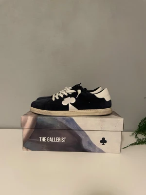  The Gallerist sneakers - Snygga blåa sneakers från The Gallerist med vit klöverdetalj på sidan och vita skosnören. Skorna har vit sula och vita detaljer vid hälen. Ovandelen är i mocka och insidan har tryckt text. Perfekta för dig som vill sticka ut med stilrena detaljer.